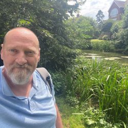 A man in a blue t-shirt standing on a river bank.